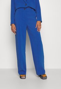 Blauw op maat gemaakte pak met wijde broekspijpen en een bijpassende jas. Kenmerkt zich door een zachte textuur, twee knopen en een strakke silhouet. Geaccentueerd met plateau schoenen.