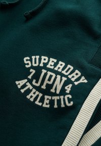 Donkergroen sweatshirt met een witte cirkelvormige print met de tekst "SUPERDAY 7JPN4 ATHLETIC." Geweven stof met geribbelde accenten.
