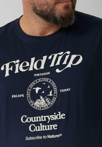 Granatowa bawełniana koszulka z białym nadrukiem graficznym. Tekst zawiera "Wycieczka" oraz "Kultura Wiejska", z okrągłym motywem przyrodniczym.