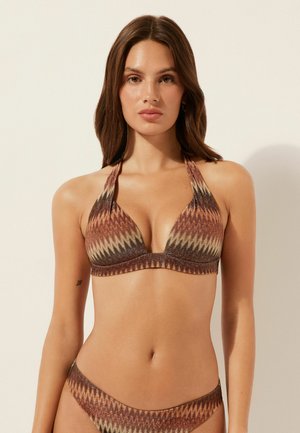 Kvinde iført en brun og beige halterneck bikini med zigzag-mønster og matchende underdel foran en offwhite baggrund.