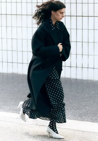 Zwarte oversized jas over een zwart stippenjurk met ruches, gecombineerd met witte stippenpanty's en witte hakken.