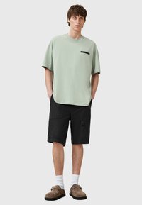 Camiseta de algodón verde con mangas cortas y detalle de bolsillo, combinada con pantalones cortos de carga negros, calcetines blancos y zapatos de slip-on marrones.
