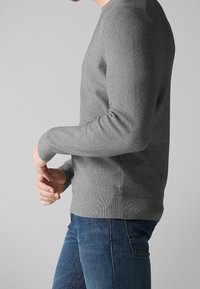 Suéter de punto gris con mangas largas, puños y dobladillo acanalados, escote redondeado. Corte casual, combinado con jeans azules. Superficie de tejido texturizada.