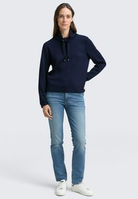 Marineblauer gerippter Pullover mit hohem Kragen, verstellbarem Zugband und langen Ärmeln, kombiniert mit hellblauen Jeans und weißen Sneakers.