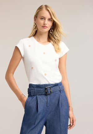 Blond vrouw met los haar draagt een witte korte mouwen top met gouden accenten, blauwe hoge taille broek met gevlochten riem en gouden oorringen.