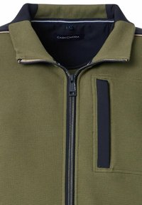 Olivgrüne Zip-Jacke aus strukturiertem Stoff mit einem schwarzen Reißverschluss und Taschenakzenten. Blau-weiße Streifen am Kragen.