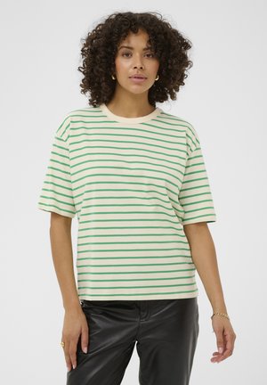 Göndör hajú nő krémszínű és zöld csíkos pólóban, fekete bőr nadrágot viselve, egyszínű fehér háttér előtt áll.