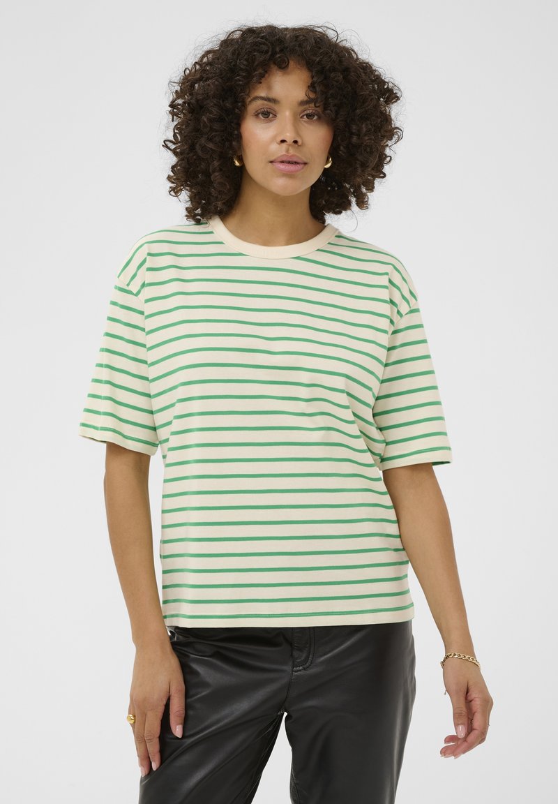 Sieviete ar cirtainiem matiem, tērpusies krēmkrāsas un zaļās svītrās T-kreklā un melnās ādas biksēs, stāvot pret vienkāršu balto fonu.