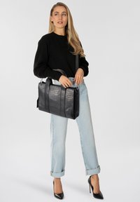 Vrouw met lang blond haar, gekleed in een zwarte trui, lichtblauwe opgerolde spijkerbroek en zwarte hoge hakken, terwijl ze een zwarte gestructureerde handtas vasthoudt.