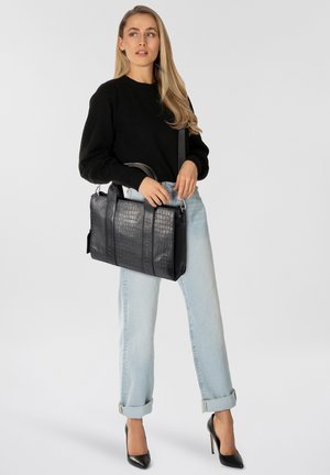 Mujer con cabello rubio largo, vistiendo un suéter negro, pantalones vaqueros azul claro arremangados y tacones altos negros, sosteniendo un bolso de mano negro texturizado.