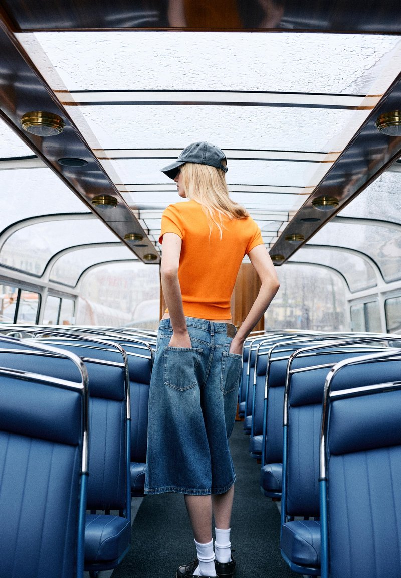 Junge Frau in orangefarbenem Hemd und Jeansrock steht in einem leeren Glasdachbus, die Hände in den hinteren Taschen, schaut zur Seite.