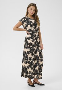 Maxiklänning i beige med svart bladmönster, korta ärmar, rund halsringning och flödande tyg. Bärs med svarta spetsskor.