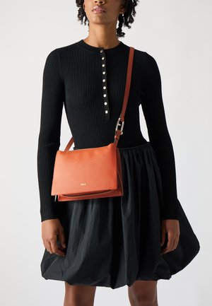 Borsa a tracolla in pelle arancione con finitura testurizzata, completata da una tracolla regolabile marrone. Il modello indossa un vestito nero a coste e una gonna.