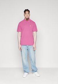 Camisa polo de algodón rosa con cuello, mangas cortas y un pequeño logo amarillo. Combinada con jeans de mezclilla azul claro y zapatillas blancas.