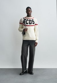 Weißer Pullover mit schwarzem und rotem Logo, kombiniert mit dunkelgrauen Jeans. Minimalistisches Design mit lockerer Passform, ergänzt durch eine kleine schwarze Tasche.
