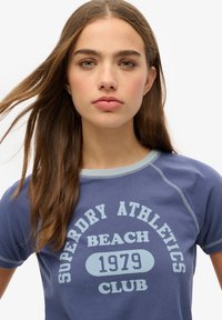 Camiseta de algodón azul con mangas cortas, que presenta un texto en color azul claro que dice "Superdry Athletics Beach Club 1979" con un diseño redondeado.