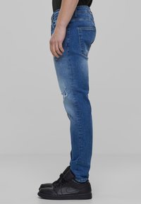 Jeans de mezclilla azul con un corte ajustado, que presentan un detalle desgastado en el muslo izquierdo y bolsillos estándar. Combinados con zapatillas altas negras.
