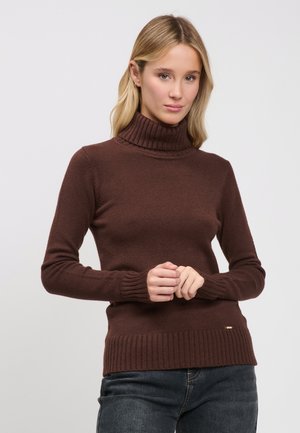 Blonde Frau trägt einen braunen gerippten Rollkragenpullover und dunkle Jeans, steht mit sanft vor dem Körper verschränkten Händen.
