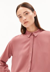 Young woman with light freckles and straight brown hair wears a dusty pink button-up shirt, posing against a plain white background.