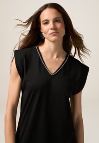 Top negro de cuello en V con mangas cortas estructuradas, que presenta una línea decorativa de diamantes de imitación a lo largo del escote. Textura de tela suave.