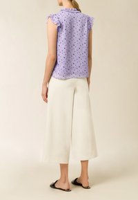 Blusa lilla con maniche a ruffle, caratterizzata da un motivo a righe con cuori blu; indossata con pantaloni bianchi a gamba larga e sandali piatti neri.