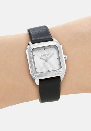 Montre carrée argentée avec cadran blanc, bracelet en cuir noir et index horaires noirs minimalistes portée au poignet, marque DKNY visible sur le cadran.