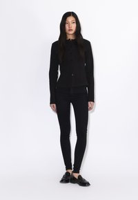 Mujer con cabello largo y oscuro, vistiendo una chaqueta negra ajustada con botones, pantalones negros ajustados y mocasines negros, de pie sobre un fondo blanco liso.