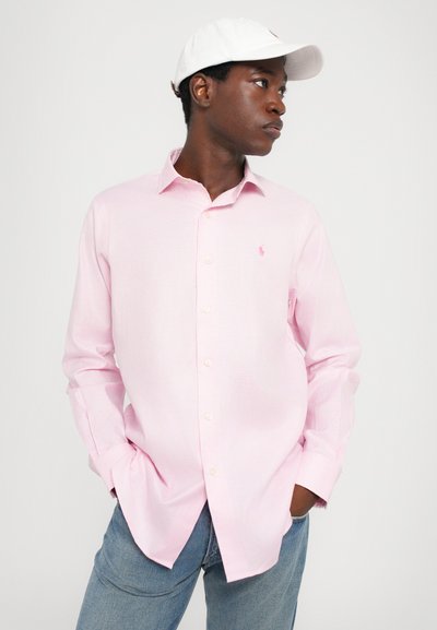 Φωτεινό ροζ καρό πουκάμισο με μακριά μανίκια, που διαθέτει ένα μικρό κεντημένο λογότυπο στο στήθος, συνδυασμένο με μπλε τζιν και μια λευκή καπέλο.