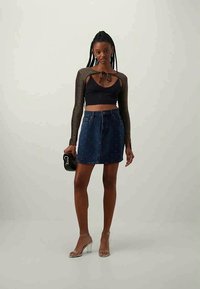 Top cropped preto com detalhe de laço na frente e mangas longas brilhantes, combinado com uma saia de ganga com tachas metálicas, usada com saltos transparentes.