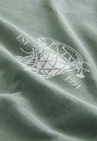 Grüner Baumwollstoff mit weicher Textur zeigt ein weißes Grafikdesign eines Basketballs und den Text "EST 1991".