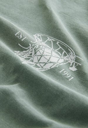 Grøn bomuldsstof med en blød tekstur har et hvidt grafisk design af en basketball og teksten "EST 1991".