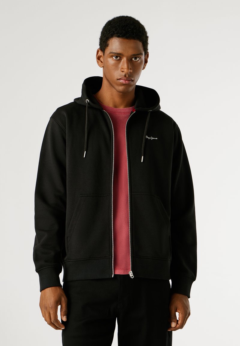 Μαύρο φούτερ με φερμουάρ και μπροστινή τσέπη, με κουκούλα που ρυθμίζεται με κορδόνι, φορεμένο πάνω από ένα κόκκινο μπλουζάκι, υφή υφάσματος και διακριτική σήμανση στο στήθος.