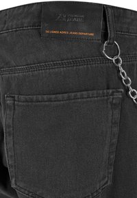 Čierne riflové nohavice s koženou nášivkou s označením "2XY PREMIUM JEANS." Obsahujú reťazový akcent pripevnený k pútku na opasok.
