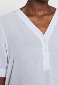 Svetle modré tričko s krátkým rukávem a výstřihem do V, vyrobené z texturované tkaniny a zdobené třemi knoflíky na výstřihu. Doplňuje ho jednoduchý lem.