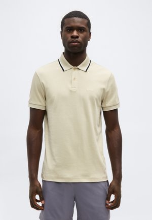 Egy férfi bézs rövid ujjú pólóingben, fekete szegéllyel díszített gallérral és szürke rövidnadrágban, egyszínű háttér előtt áll.