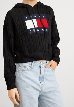 Person wearing black cable-knit cropped hoodie with Tommy Jeans logo and light blue high-waist jeans, one hand in pocket.