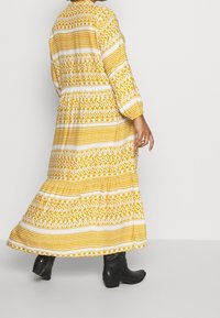 Person som bär en lång gul och vit mönstrad klänning och svarta ankelboots, går med en hand vid sin sida och visar en silverring.