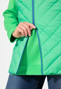 Chaqueta verde con cremallera y textura acolchada, con un patrón de chevron, que presenta una cremallera azul. Se lleva sobre un suéter de cuello redondo verde sólido.