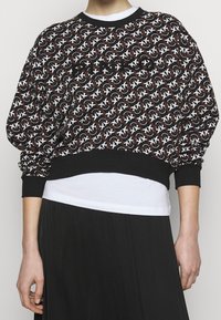 Czarna i biała bluza z wzorem, z prążkowanym okrągłym dekoltem i elastycznymi mankietami, noszona na białej koszuli, zestawiona z czarną spódnicą.