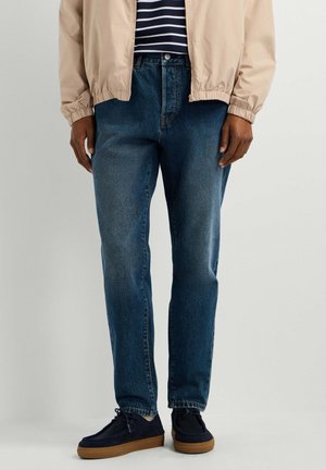 Mann iført blå jeans, marineblå sko med brune såler, beige jakke og marineblå og hvit stripete skjorte, stående på lys bakgrunn.