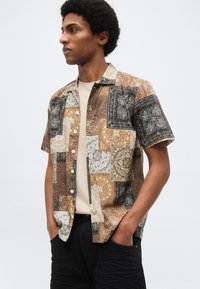 Άνδρας με αφρό κούρεμα που φοράει κουμπωτό πουκάμισο με κοντά μανίκια και ραμμένα σχέδια πάνω από μπεζ μπλουζάκι, με μαύρο παντελόνι, χέρια στις τσέπες.