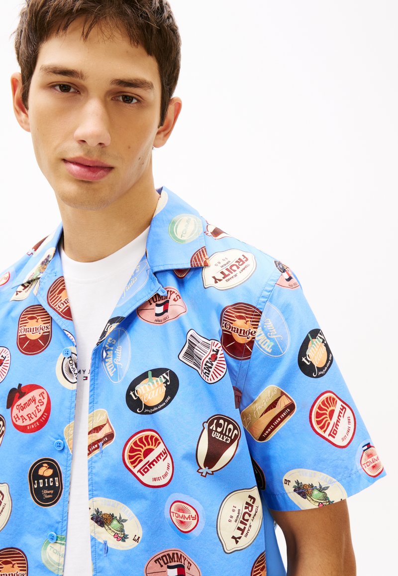 Jeune homme portant une chemise à manches courtes bleue avec des patchs colorés de style vintage représentant des aliments et des logos de marques par-dessus un T-shirt blanc.