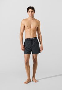 Homem sem camisa, descalço, a usar calções de banho pretos com a cinta da marca Karl Lagerfeld visível, em frente a um fundo claro.