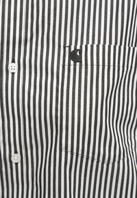 Gestreifte schwarz-weiße Button-Up-Bluse mit einer Brusttasche und einem gestickten Logo. Der Stoff wirkt glatt und hat eine leichte Textur.