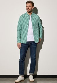 Green checked button-up shirt with long sleeves, paired with a white T-shirt, blue jeans, and white sneakers. Smooth textures throughout.