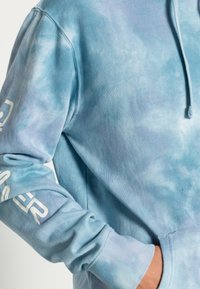 Light blue tie-dye hoodie, cotton material, soft texture, kangaroo pocket, drawstring hood, white logo on sleeve, relaxed fit.