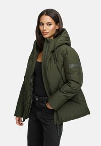 Femme aux cheveux lisses et foncés portant une doudoune olive ouverte, un haut noir et un pantalon noir, se tenant avec une main dans la poche.