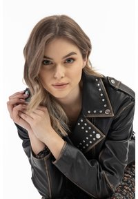 Zwarte leren jack met zilveren studs, asymmetrische kraag en ritsdetails. Model met golvend haar poseert met handen dichtbij het gezicht.