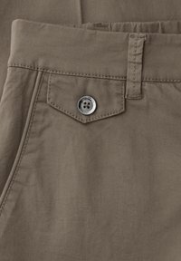 Gros plan sur la poche d’un pantalon kaki avec rabat maintenu par un bouton métallique à quatre trous et passants pour ceinture à la taille.