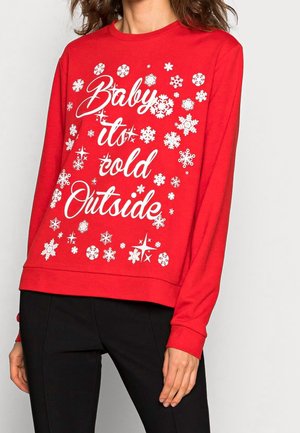 Czerwona bluza z długim rękawem z białymi płatkami śniegu i napisem "Baby it's cold Outside" noszona z czarnymi spodniami.